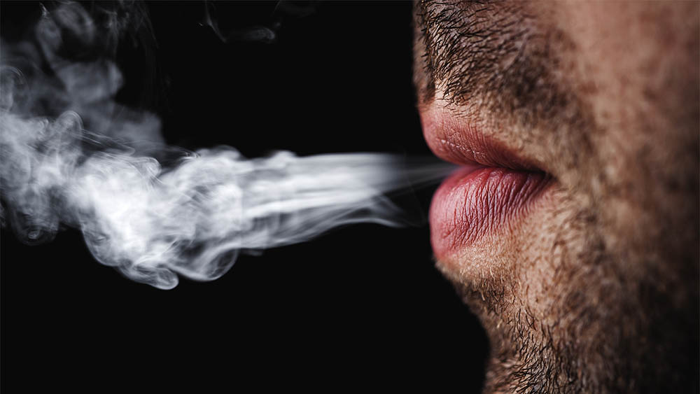 Close em uma boca de homem de perfil exalando fumaça de cigarro em um fundo preto.