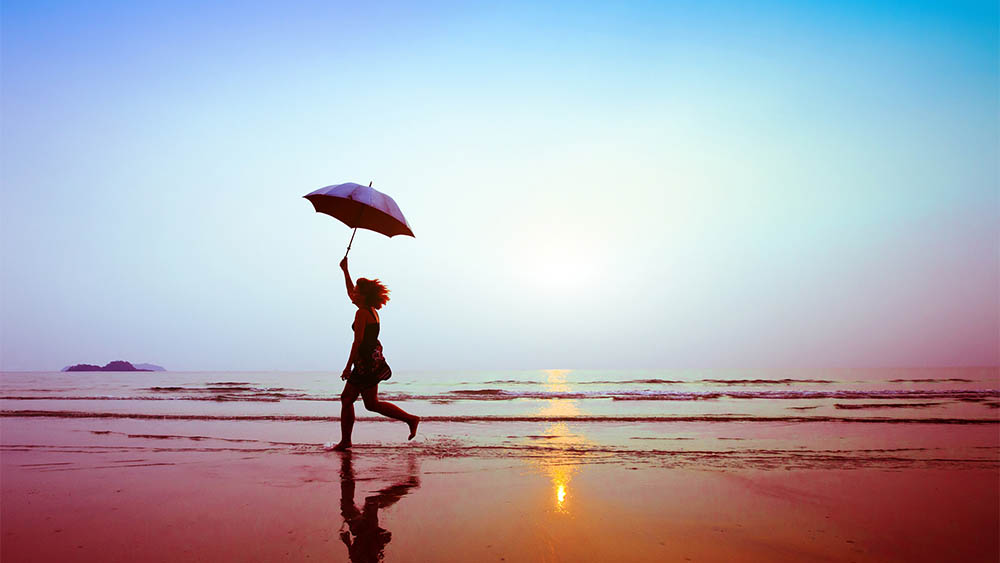 Mulher correndo na praia segurando guarda-chuva.