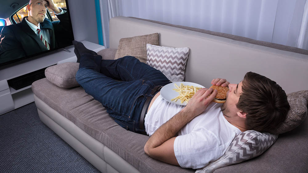 Homem jovem deitado em sofá comendo hambúrguer e assistindo a televisão.